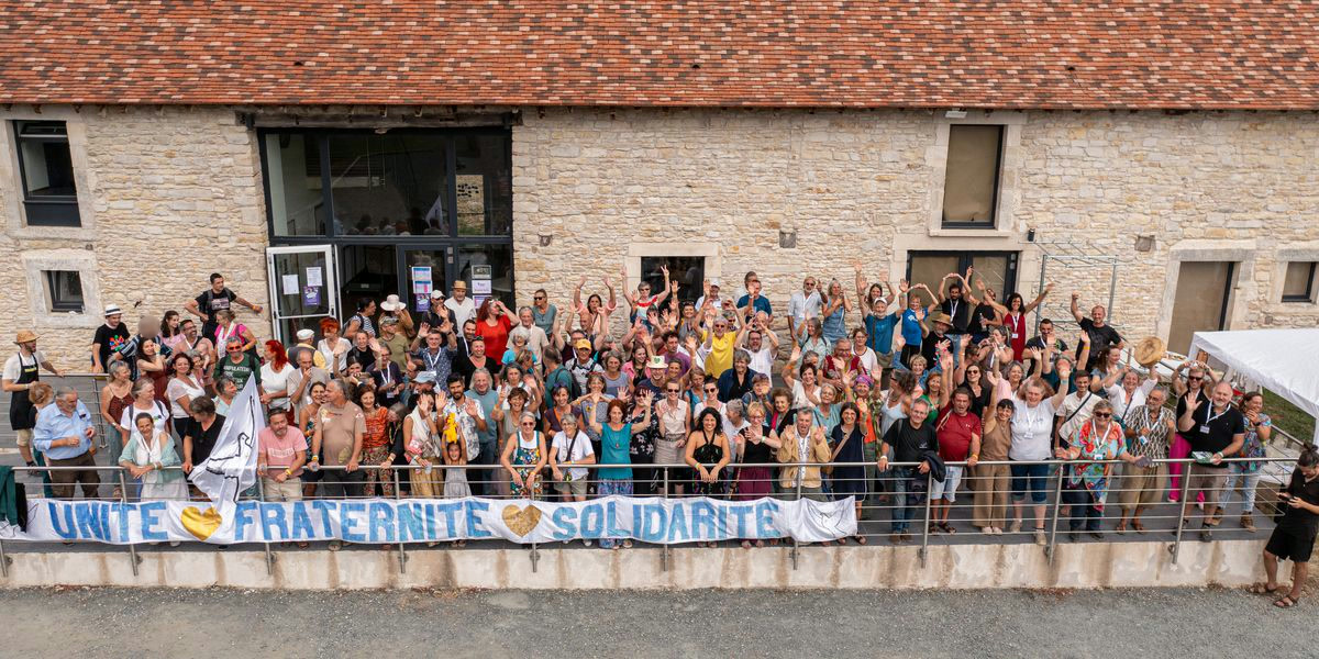 Les participants d'Émancip'Actions 2026 réunis devant un bâtiment en pierre, derrière une banderole « Unité, Fraternité, Solidarité ». Beaucoup lèvent les bras.